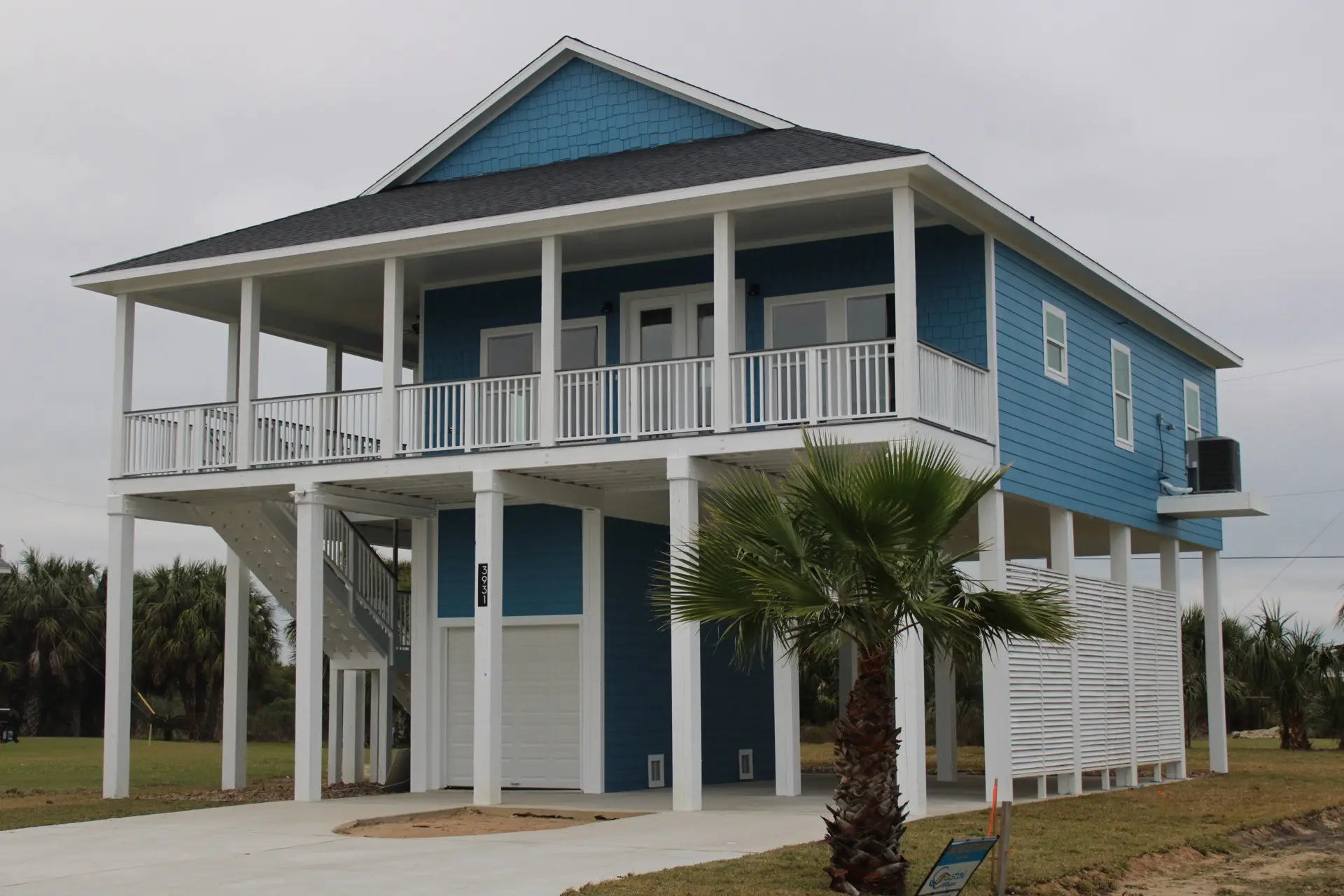 Front exterior of 31 Laguna on Tiki Island featuring bold blue siding with white columns and railing, a large wraparound covered porch with ceiling fans, and palm tree landscaping