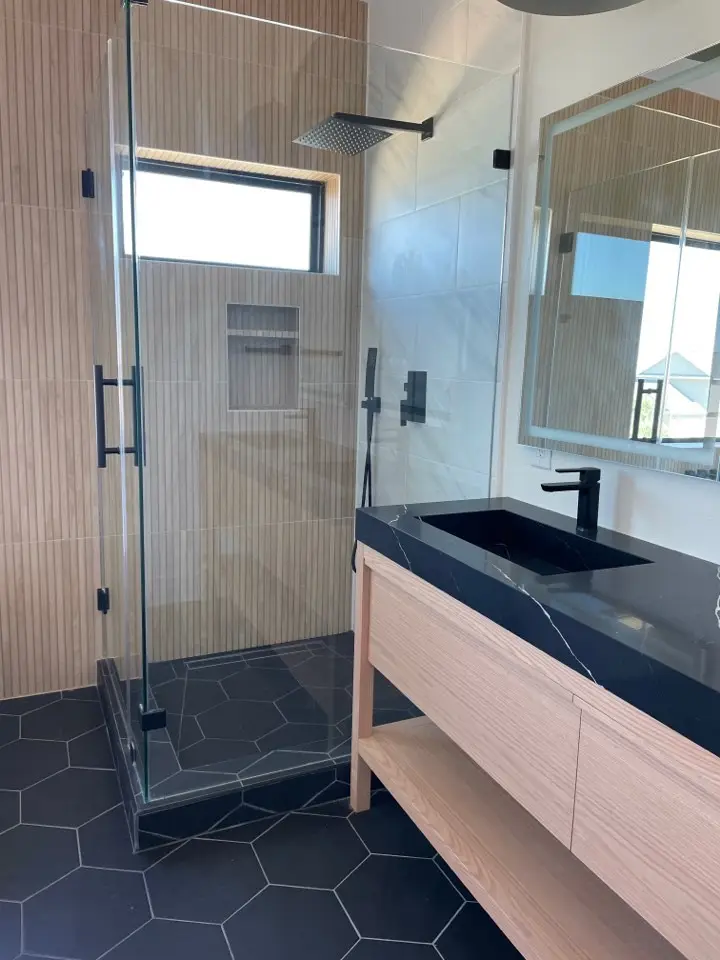 Primary bathroom vanity with dual sinks, large rectangular mirror, contemporary wall sconces, and dark hexagon floor tile extending beneath the tub